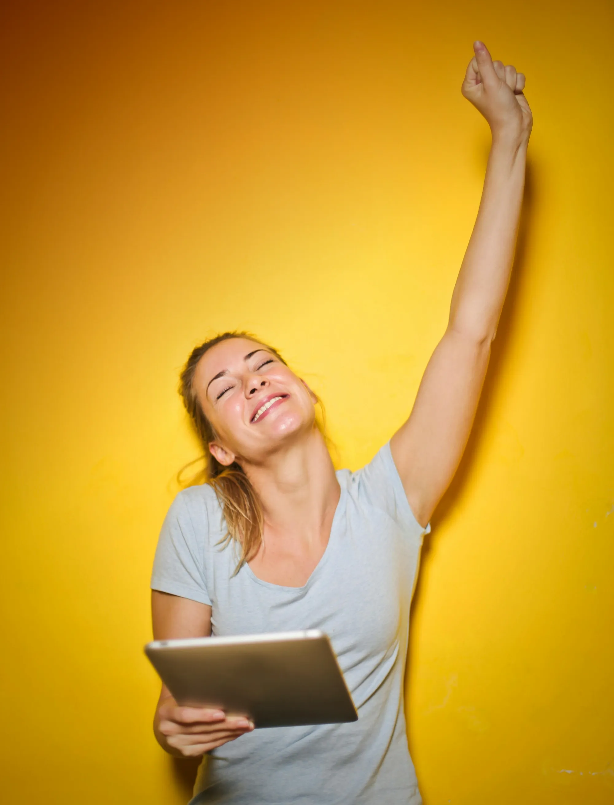 woman with arm outstretched while holding a digital tablet and smiling on yellow background